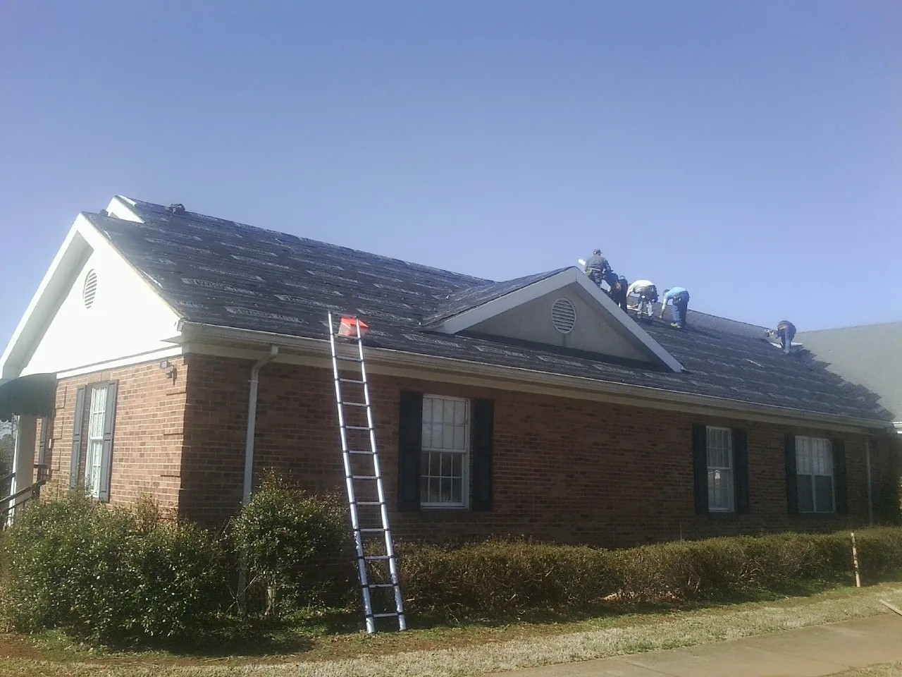 Crew tearing shingles off a brick ranch roof with a ladder set — Metal Roof Repair work by our crew in Harmans